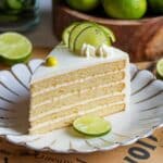 A beautifully sliced piece of layered Key Lime Cake Recipe topped with lime slices and creamy frosting, served on a decorative plate with fresh limes in the background.