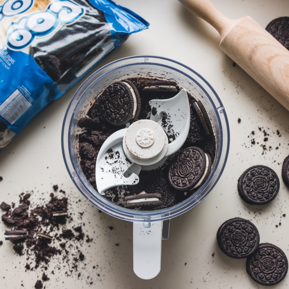 A playful kitchen scene with Oreos in a food processor, crushed crumbs on the counter, and a rolling pin nearby, capturing the process of preparing the dirt layer for the Oreo Dirt Cake Recipe.