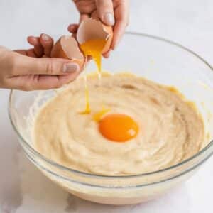 A close-up of a person cracking an egg into a mixing bowl filled with a smooth batter for a Cream Cheese Brownies Recipe. The rich, creamy mixture is being prepared for the cheesecake swirl, ensuring a luscious and decadent texture in the final baked brownies.