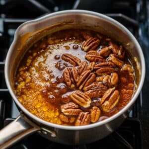 A saucepan filled with a bubbling caramel pecan topping for a Pecan Pie Cheesecake Recipe. The mixture of butter, sugar, heavy cream, and corn syrup thickens as it simmers, with toasted pecans and vanilla added for a rich, nutty flavor. A perfect topping for a decadent cheesecake dessert.