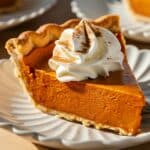 A slice of Pumpkin Pie with Condensed Milk on a decorative white plate, featuring a smooth, creamy filling with a deep orange hue and a flaky golden crust. Topped with a swirl of whipped cream and a light dusting of cinnamon, this classic fall dessert is captured in warm, natural lighting, highlighting its rich texture and inviting appeal.