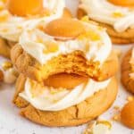 Soft and chewy Banana Pudding Cookies topped with creamy frosting, a vanilla wafer, and crushed wafer pieces. These golden-brown cookies are made with banana pudding mix, white chocolate chips, and vanilla wafers for a rich, sweet flavor. A bite taken out of one reveals a moist, cake-like texture. This Banana Pudding Cookie Recipe is a delicious twist on the classic banana pudding dessert.