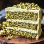 Homemade pistachio cake slice with three green layers, creamy white frosting, and chopped roasted pistachios on top, served on a rustic wooden board.