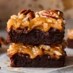 Close-up of two stacked homemade German chocolate brownies with a fudgy chocolate base and a thick, glossy coconut-pecan frosting. The caramel-colored frosting is rich and gooey, topped with toasted coconut flakes and crunchy pecan halves. The brownies have a moist texture with melted chocolate chips visible, and the frosting slightly drips over the edges. Warm, natural lighting enhances the rich textures, creating a decadent and homemade feel.