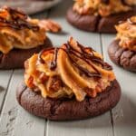 A batch of homemade German chocolate cookies with a thick, fudgy chocolate base. Each cookie is topped with a generous portion of golden-brown coconut-pecan caramel frosting, which has visible coconut shreds and crunchy pecan pieces. The frosting appears rich and slightly textured, with a soft and moist consistency.