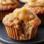 Homemade applesauce muffins with a bite taken out, showing its moist, golden crumb and topped with apple chunks, on a white plate with blurred muffins in the background.