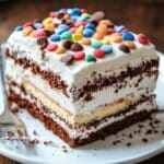Homemade ice cream sandwich cake slice with layers of chocolate cake, vanilla ice cream, and whipped topping, garnished with colorful candies and chocolate chips on a white plate.