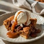 A homemade pecan pie cobbler slice on a textured off-white plate, topped with a scoop of vanilla ice cream and drizzled with caramel sauce. The golden, flaky crust reveals a gooey, caramelized pecan filling with toasted pecans on top. Sunlight casts warm highlights, with a blurred baking dish and linen cloth in the background, enhancing the rustic, homemade feel.
