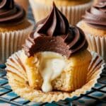 A homemade Boston Cream Cupcakes with a bite taken out, revealing creamy vanilla custard oozing from the soft, golden sponge. Topped with a rich, glossy chocolate swirl, the cupcake sits in a slightly crinkled white liner on a black cooling rack.