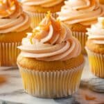 Close-up view of homemade orange cupcakes topped with fluffy, swirl-shaped pale-orange buttercream frosting and garnished with fresh orange zest. The cupcakes are baked to a perfect golden color and presented neatly in white paper cupcake liners. The scene is set on a marble surface, highlighting a fresh, inviting, and authentic homemade appearance.