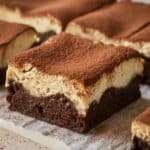 Homemade tiramisu brownies with a fudgy chocolate base and creamy mascarpone layer, dusted with cocoa powder. Cut into squares on parchment paper, with a rich, moist texture and a rustic homemade feel.