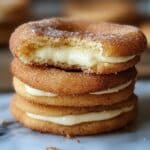 Stack of churro cheesecake cookies with cinnamon-sugar coating and creamy filling, one with a bite showing soft, moist inside.