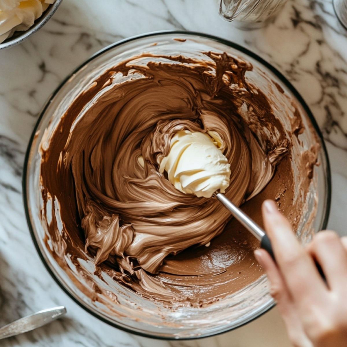 A close-up of a glass mixing bowl with a creamy chocolate mixture, as softened butter is being stirred in with a spatula. The mixture is smooth and fluffy, with soft natural light highlighting the texture on the marble countertop.
