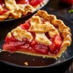 A slice of homemade strawberry rhubarb pie with a golden, flaky lattice crust and glossy, juicy fruit filling on a black plate. The rustic texture and vibrant colors give it a fresh, real-baked look.