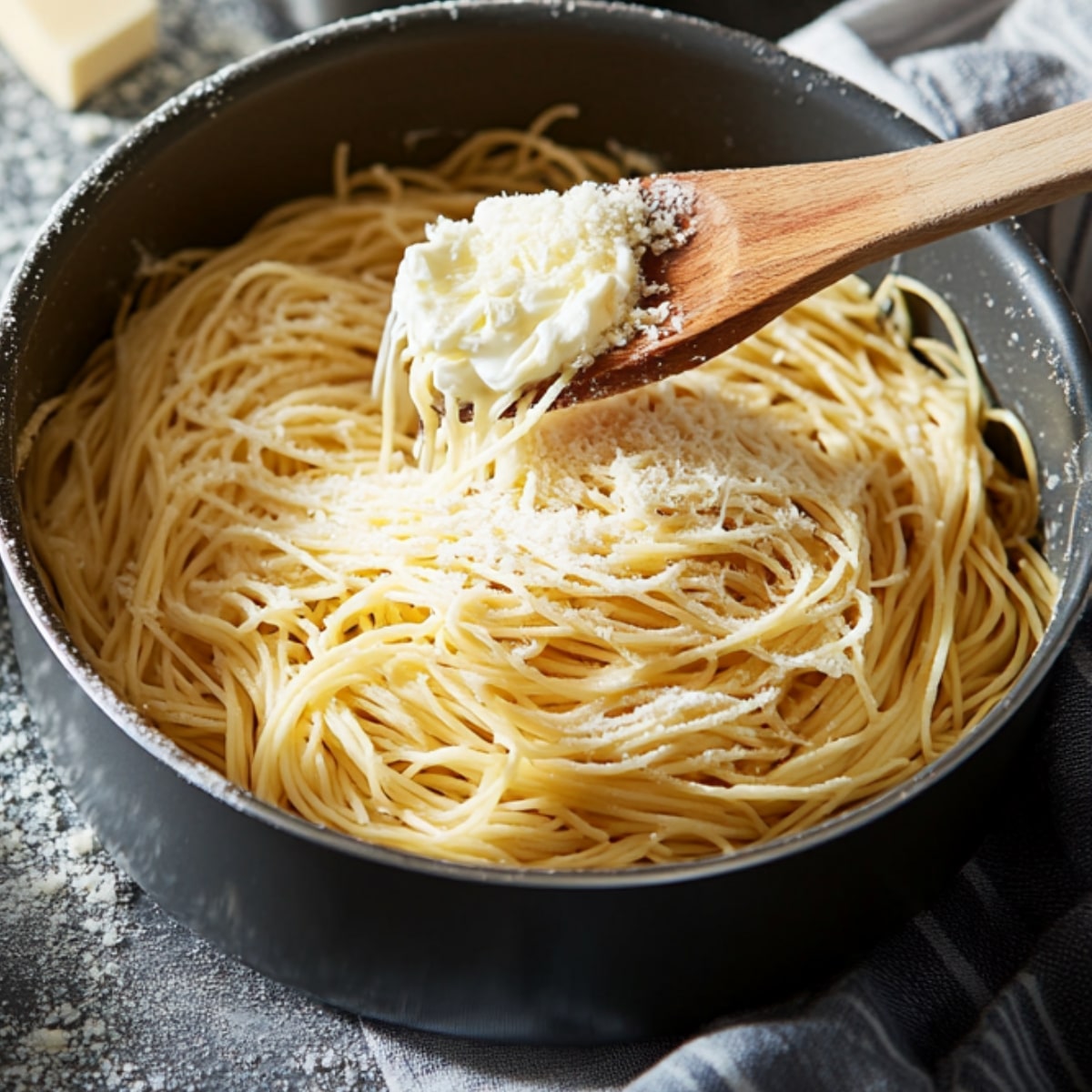 Cooked spaghetti in a black pot being tossed with butter and grated Parmesan cheese, with a wooden spoon lifting the pasta. The scene has a cozy, homemade kitchen feel.