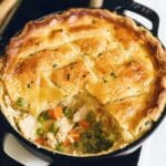 Homemade chicken pot pie in a black skillet with a golden, flaky crust, cut open to reveal a creamy filling of chicken, peas, and carrots, topped with parsley and black pepper.