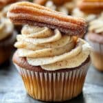 A close-up of a homemade churro cupcake with a golden-brown base, topped with cinnamon buttercream frosting and a crispy churro stick dusted with powdered sugar. The white cupcake wrapper and cinnamon sugar finish give it a warm, homemade feel.