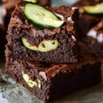 Homemade fudgy zucchini brownies with glossy, cracked tops and fresh zucchini slices on top. The brownies have a rich, dense texture, with a slight crisp on the edges, resting on parchment paper for a homemade feel.