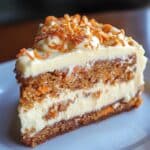 A thick slice of homemade carrot cake cheesecake on a white plate, with two moist carrot cake layers filled with shreds of carrot, a creamy cheesecake center, and topped with swirls of cream cheese frosting and caramelized carrot garnish.