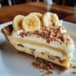 Homemade banana cream pie slice with fresh banana slices, creamy custard, whipped topping, and a graham cracker crust, served on a white plate with a cozy, rustic look.