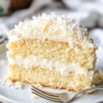 A slice of homemade coconut cake with three layers of fluffy vanilla sponge, creamy coconut frosting, and shredded coconut on top, served on a white plate with a fork beside it.
