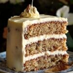 A homemade slice of Hummingbird Cake with three moist layers filled with banana, pineapple, and pecans, frosted with creamy cream cheese icing, served on a vintage floral plate with a gold fork.