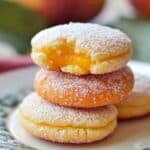 Stack of five homemade peach cookies on a white plate, sugar-coated with golden edges and glossy peach filling in the center, softly lit by natural daylight with a blurred green background.
