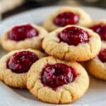Homemade raspberry thumbprint cookies on a white embossed plate, golden and slightly cracked, each filled with glossy, seed-speckled raspberry jam, sitting on a rustic wooden table.