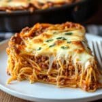 A slice of homemade spaghetti pie Recipe on a white plate, featuring layers of spaghetti, meaty tomato sauce, and melted mozzarella cheese, garnished with fresh parsley. The dish has a rich, inviting look with a rustic, homemade feel.