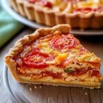 Slice of homemade tomato pie with flaky crust, roasted tomatoes, melted cheese, and creamy filling on a rustic plate, with the full pie in the background.