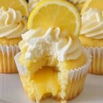 A homemade lemon cupcake with fluffy yellow cake, topped with swirled white frosting and a lemon slice. A bite reveals a gooey lemon curd filling oozing from the center, with more cupcakes blurred in the background.
