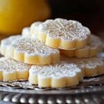 Close-up of homemade lemon meltaway cookies, stacked on a vintage silver tray. The cookies are shaped like flowers, lightly dusted with powdered sugar, and have a soft yellow color. A blurred lemon is visible in the background, adding a fresh touch to the rustic, cozy scene.