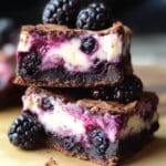 Homemade blackberry cheesecake brownies with fudgy chocolate layers, creamy cheesecake swirls, and baked blackberries, stacked on parchment with fresh berries on top.