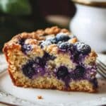 Close-up of a homemade blueberry coffee cake slice on a white plate with a silver border. The cake has a golden-brown, crumbly top, with burst blueberries and a dusting of powdered sugar. The soft, moist interior reveals scattered blueberries, with a blurred porcelain item in the background, adding a cozy, homemade feel.