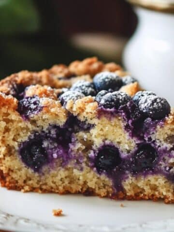 Close-up of a homemade blueberry coffee cake slice on a white plate with a silver border. The cake has a golden-brown, crumbly top, with burst blueberries and a dusting of powdered sugar. The soft, moist interior reveals scattered blueberries, with a blurred porcelain item in the background, adding a cozy, homemade feel.