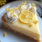 A slice of homemade no bake lemon pie with whipped cream, lemon slice, and yellow flowers on a ceramic plate.