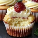 Homemade cheesecake cupcakes with creamy frosting, a strawberry on top, and a bite showing the soft vanilla cake inside.
