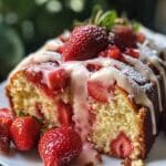 A thick slice of moist strawberry pound cake topped with fresh strawberries, a light pink glaze, and powdered sugar. Juicy strawberries are baked into the golden crumb, and the cake sits on a white plate with natural light in the background.