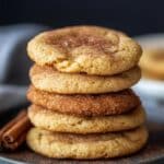 Stack of freshly baked snickerdoodle cookies with a cinnamon-sugar coating, one with a bite taken out, resting on a gray plate with a cinnamon stick nearby. The soft, chewy texture is highlighted against a blurred background.
