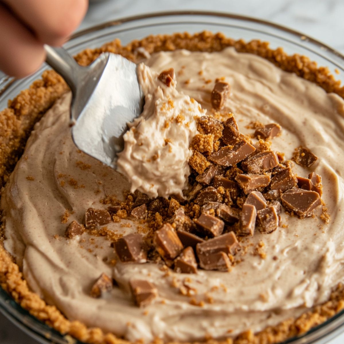 Close-up of Butterfinger Pie being filled with creamy candy mixture and topped with crushed Butterfinger pieces in a graham cracker crust.