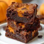 Fudgy pumpkin brownies stacked on a white plate, with rich chocolate layers and swirls of pumpkin. Crackly tops, gooey texture, and mini pumpkins blurred in the background for a cozy fall feel.
