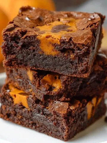 Fudgy pumpkin brownies stacked on a white plate, with rich chocolate layers and swirls of pumpkin. Crackly tops, gooey texture, and mini pumpkins blurred in the background for a cozy fall feel.