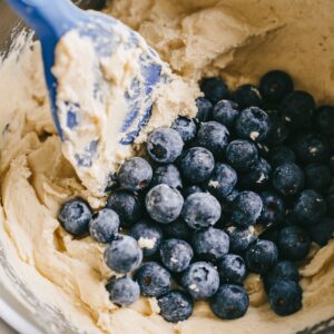 Fresh blueberries being gently folded into thick cookie dough with a blue spatula in a mixing bowl.
