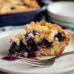 A homemade slice of blueberry dump cake topped with whipped cream, fresh blueberries, and powdered sugar, served on a white plate with a fork.