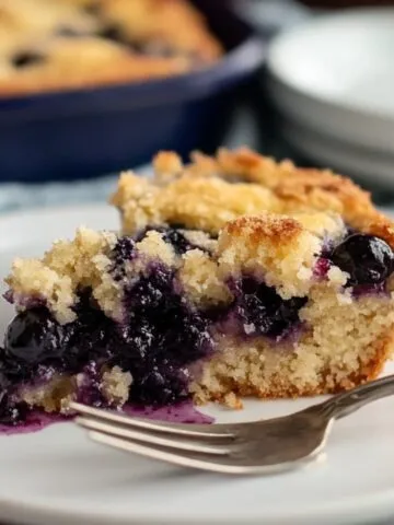 A homemade slice of blueberry dump cake topped with whipped cream, fresh blueberries, and powdered sugar, served on a white plate with a fork.