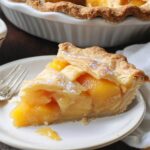 Slice of homemade canned peach pie with golden lattice crust and juicy peaches on a white plate, next to a whipped cream bowl and the full pie in a ceramic dish.