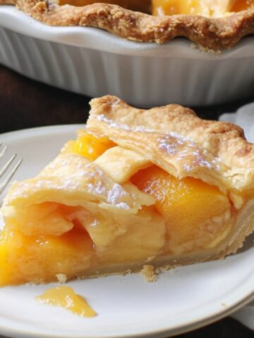 Slice of homemade canned peach pie with golden lattice crust and juicy peaches on a white plate, next to a whipped cream bowl and the full pie in a ceramic dish.