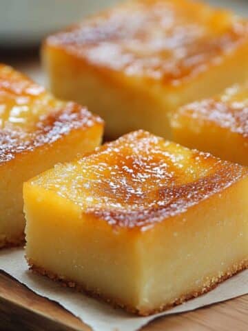 Close-up of freshly baked homemade cassava cake recipe , sliced into neat golden-brown squares and arranged on a piece of parchment paper over a wooden board. Each slice has a glossy, caramelized top with a slightly crisp edge and a moist, dense interior that reveals the rich yellow hue of the cassava base.