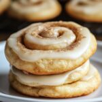 Homemade cinnamon roll cookies stacked on a plate, swirled with icing and dusted with cinnamon sugar, with more cookies cooling in the background.
