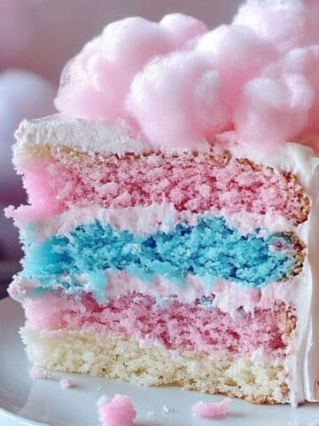 Homemade cotton candy cake slice with pink, blue, and white layers, fluffy frosting, and a pink cotton candy topping, served on a white plate with candy pearls.